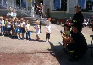 Widok na grupę dzieci i dziewczynkę, która leje wodą z węża strażackiego.