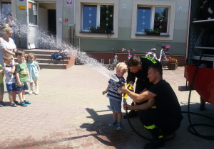 Widok na grupę dzieci i chłopca, który leje wodą z węża strażackiego.