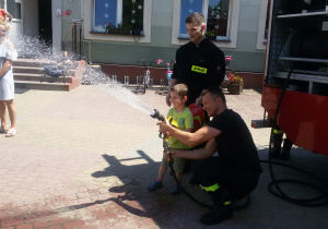 Widok na strażaków i chłopca, który leje wodą z węża strażackiego.