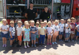 Dzieci pozują do zdjęcia grupowego ze strażakami. W tle widać samochód strażacki.