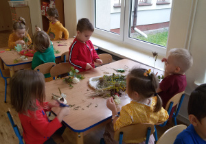 Widok na dzieci siedzące przy stolikach i wykonujące prace plastyczne z tworzywa przyrodniczego i masy solnej.