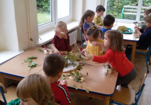 Widok na dzieci, które siedzą przy stolikach i wykonują wazoniki z jesiennymi bukietami.
