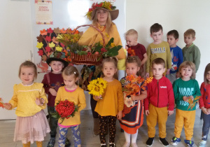 Maluchy pozują do zdjęcia grupowego z Panią Jesienią, która trzyma w ręku kosz z jesiennymi darami. 4 dzieci trzyma w rękach kolorowe liście, Marysia trzyma bukiet słoneczników, Nadia bukiet jarzębiny.