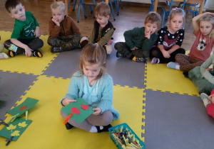Widok na siedzące w kręgu dzieci oraz sadzonkę sosny. W środku kręgu siedzi Marysia trzymając w rękach zielony kartonik z 2 listkami i 2 klamerkami.