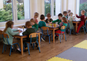 Widok na siedzące przy 3 stolikach dzieci, które naklejają bibułkowe listki na „zielone korony drzew”.