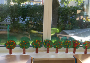 Widok na wykonane przez dzieci jesienne drzewka, które stoją na parapecie okna. Za oknem widać zieleń i na budynki.