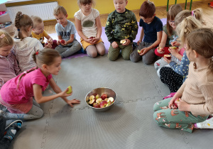 Dzieci siedzą w kole na dywanie i oglądają połówki jabłek. Na środku stoi miska z jabłkami.