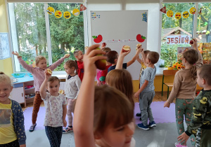 Dzieci poruszają się przy muzyce trzymając połówki jabłek w dłoni, uniesioną w górze.
