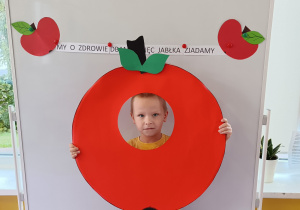 Dziecko pozuje do zdjęcia z głową umieszczoną w kształcie tekturowego jabłka. W tle napis „My o zdrowie dbamy więc jabłka zjadamy”.