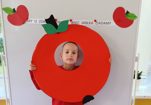 Dziecko pozuje do zdjęcia z głową umieszczoną w kształcie tekturowego jabłka. W tle napis „My o zdrowie dbamy więc jabłka zjadamy”.