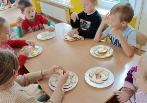 Dzieci siedzą przy stoliku i układają na talerzu kompozycję z pokrojonych w plastry jabłek.