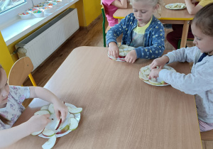 Dzieci siedzą przy stoliku i układają na talerzu kompozycję z pokrojonych w plastry jabłek.