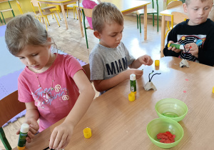 Troje dzieci siedzi przy stoliku i tworzy przestrzenne „drzewko jabłoni” z tekturki i drucika kreatywnego. Przyklejają papierowe czerwone jabłuszka.