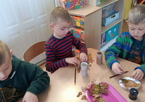 3 chłopców siedząc przy stole tworzy jesienne drzewo z naturalnych liści i rolki.