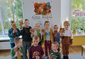 Grupa dzieci pozuje do zdjęcia trzymając w ręku samodzielnie zrobione przestrzenne drzewa. W tle tablica magnetyczna z obrazem „Jesień w parku” oraz napis „Dzień Drzewa”.