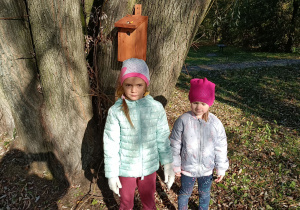Dwie dziewczynki pozują do zdjęcia przy drzewie, na którym widać budkę dla ptaków.