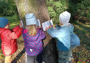Troje dzieci stoi przy drzewie i kalkuje korę drzewa przy użyciu białej kartki i kredki.