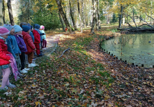Grupa dzieci stoją przy stawie i obserwują pływające kaczki.