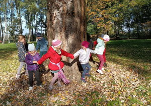 Dzieci ustawione w kole krążą wokół pnia drzewa.