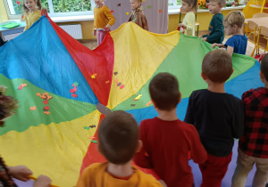 Dzieci stoją w kole, trzymają chustę animacyjną. Na chuście rozrzucone kolorowe listki z papieru.