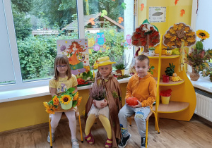 Troje dzieci siedzi na krzesłach przed „kącikiem przyrody” z dekoracją jesienną. W rękach trzymają rekwizyty: słoneczniki, wrzos, dynia.