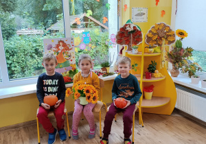 Troje dzieci siedzi na krzesłach przed „kącikiem przyrody” z dekoracją jesienną. W rękach trzymają rekwizyty: słoneczniki, dynia.