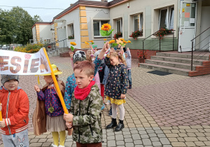 Dzieci stoją przed budynkiem przedszkola w parach. Trzymają w rękach rekwizyty: słonecznik, kolorowy listek z papieru. Pierwsza para trzyma transparent z napisem „Jesień”.