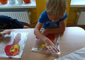 Dwoje dzieci siedzi przy stoliku. Rozprowadzają farbę(czerwona, żółta, pomarańczowa, brązowa) palcem po folii z narysowanym schematem drzewa.