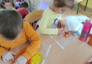 Dwoje dzieci siedzi przy stoliku. Rozprowadzają farbę(czerwona, żółta, pomarańczowa, brązowa) palcem po folii z narysowanym schematem drzewa.
