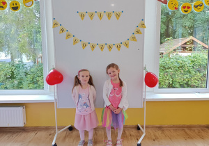Dwie dziewczynki pozują do zdjęcia. W tle tablica magnetyczna z napisem „Dzień Przedszkolaka” udekorowana czerwonymi balonami w kształcie serca.