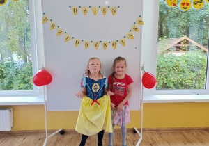 Dwie dziewczynki pozują do zdjęcia. W tle tablica magnetyczna z napisem „Dzień Przedszkolaka” udekorowana czerwonymi balonami w kształcie serca.