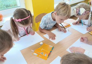 Dzieci siedzą przy stoliku i rysują swój portret trzymając mazak w ustach.