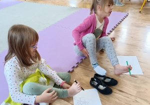 Dzieci siedzą na macie piankowej i rysują swój portret trzymając mazak stopami.