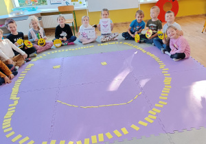 Dzieci siedzą w półkolu na dywanie trzymając w rękach symbole „uśmiechniętych buziek”. Na dywanie ułożona buźka z żółtych papierowych prostokątów.