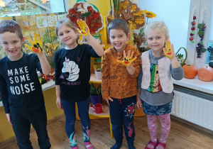 2 dziewczynki i 2 chłopców pozuje do zdjęcia z ręką uniesioną do góry. Dłonie wymalowane na żółto z symbolem buźki. W tle jesienna dekoracja.