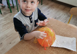 Chłopiec siedzi przy stole i owija balon włóczką tworząc dynie.