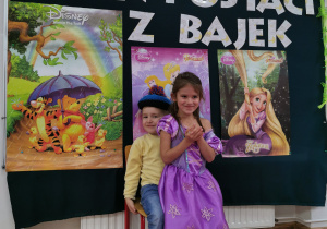Chłopiec trzyma dziewczynkę na kolanach przebrani za postacie z bajek. Tłem zdjęcia jest tablica z plakatami postaci bajkowych.