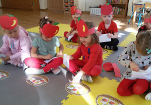 Widok na siedzące w dwóch rzędach dzieci. Przed nimi leżą papierowe koszyczki z jabłkami.
