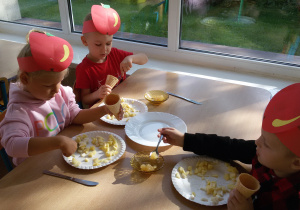 Widok na troje dzieci siedzących przy stoliku, które przygotowują deser z jabłek.