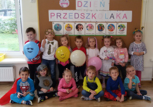 Maluchy pozują do zdjęcia grupowego na tle tablicy z napisem: Dzień Przedszkolaka. 5 dzieci trzyma kolorowe baloniki.