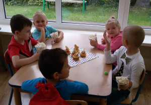Widok na siedzące przy stoliku dzieci, które jedzą popcorn. Na środku stolika leży kolorowa serwetka, a na niej babeczki z kremem.