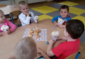 Widok na siedzące przy stoliku dzieci, które jedzą popcorn. Na środku stolika leży kolorowa serwetka, a na niej babeczki z kremem.