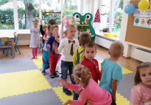 Widok na salę przedszkolną i dzieci stojące jedno za drugim w rozkroku.