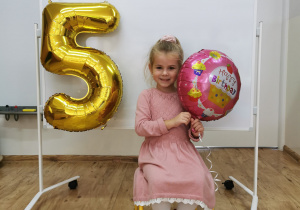 Dziewczynka pozuje do zdjęcia trzymając balon urodzinowy.