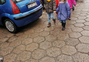 Grupa dzieci ubrana w ciepłe kurtki oraz czapki w parach kierują się w stronę autokaru.