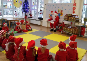 Widok na grupę siedzących w kręgu dzieci w czerwonych strojach i „mikołajkowych czapeczkach”. Przed nimi na małym krzesełku siedzi Święty Mikołaj. W tle świąteczna dekoracja.