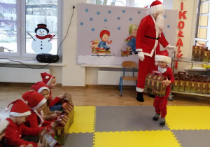Widok na salę przedszkolną, dzieci oraz Świętego Mikołaja i chłopca, który odbiera od niego prezent.