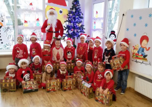 Maluchy w czerwonych strojach, z prezentami pozują do zdjęcia grupowego ze Świętym Mikołajem.
