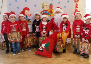 Chłopcy w „mikołajkowych strojach” pozują do zdjęcia ze Świętym Mikołajem. Każdy z chłopców trzyma w rękach „domek”- prezent od Mikołaja.