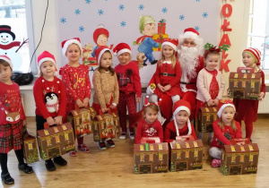 Dziewczynki w „mikołajkowych strojach” pozują do zdjęcia ze Świętym Mikołajem. Każda z nich trzyma „domek”- prezent od Mikołaja.